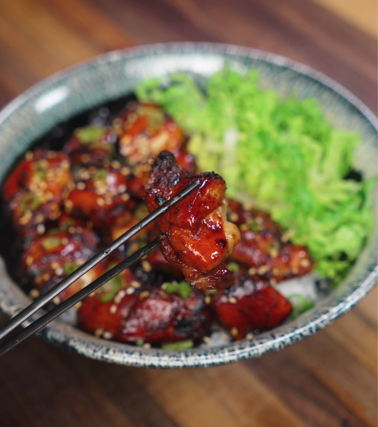 A juicy piece of garlic soy glazed chicken is held up with chopsticks over a bowl filled with caramelized chicken thighs, fluffy rice, and fresh shredded lettuce. The glossy, sticky glaze clings to the chicken, highlighting its crispy edges and tender inside, making the bite look irresistible and packed with flavor.
