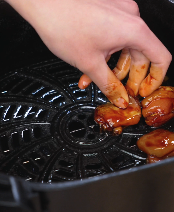 A hand carefully places marinated chicken thighs into an air fryer basket, spacing them apart for even cooking. The chicken is already coated in a glossy garlic soy glaze, showing the first step in achieving the crispy and sticky finish of the final dish.