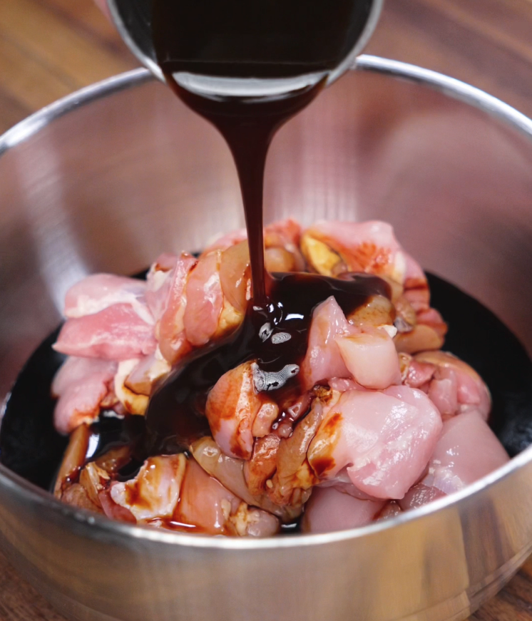 Thick garlic soy marinade is poured from a metal bowl onto bite-sized raw chicken thighs, ready to be marinated. The glossy dark sauce coats the meat, preparing it for a flavorful soak that infuses the chicken with sweet, savory, and garlicky depth before cooking.