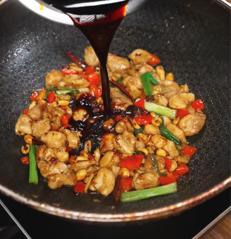 A dark, glossy sauce is poured over a pan of stir-fried chicken, peanuts, red peppers, and green onions. The rich sauce brings together the sweet, spicy, and tangy flavors that define kung pao chicken.