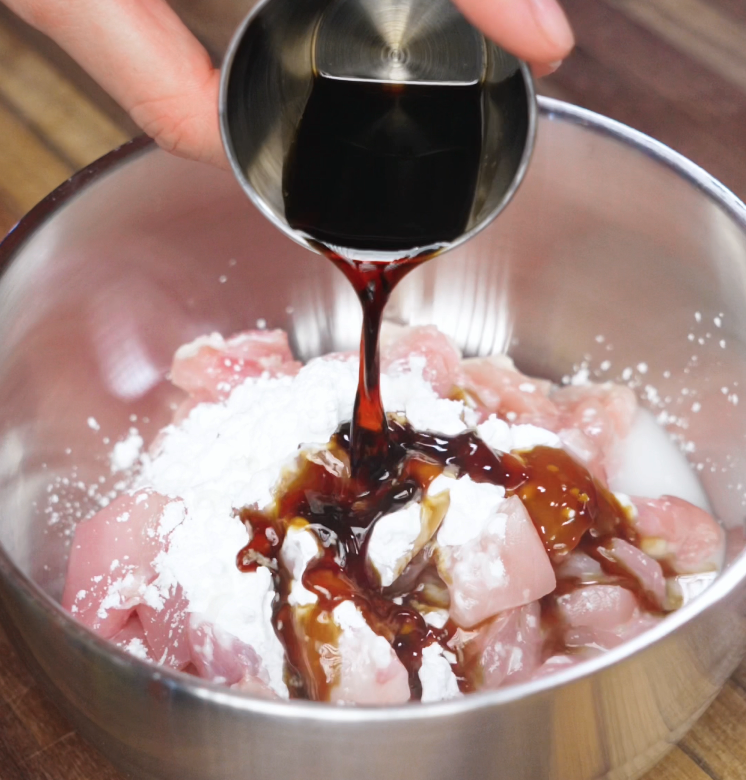 Dark soy sauce is being poured over raw chicken pieces coated in cornstarch and milk inside a mixing bowl. This marinade adds depth and color to the chicken before it’s stir-fried for kung pao chicken.