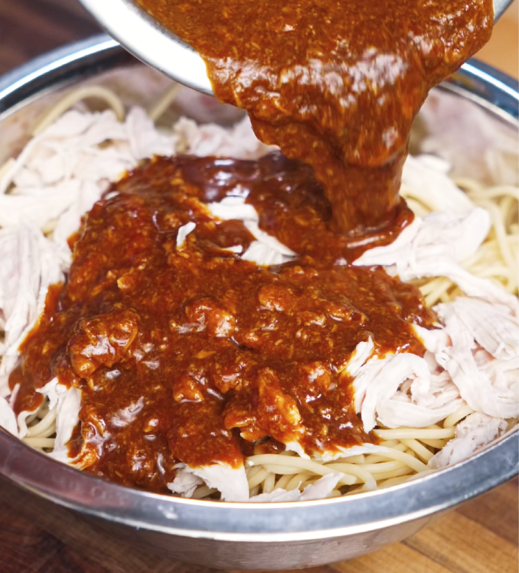 A rich, dark peanut butter-based sauce being poured over shredded poached chicken and al dente noodles in a large mixing bowl. The glossy sauce blends sweet, salty, and umami flavors that tie this Asian-inspired noodle dish together.