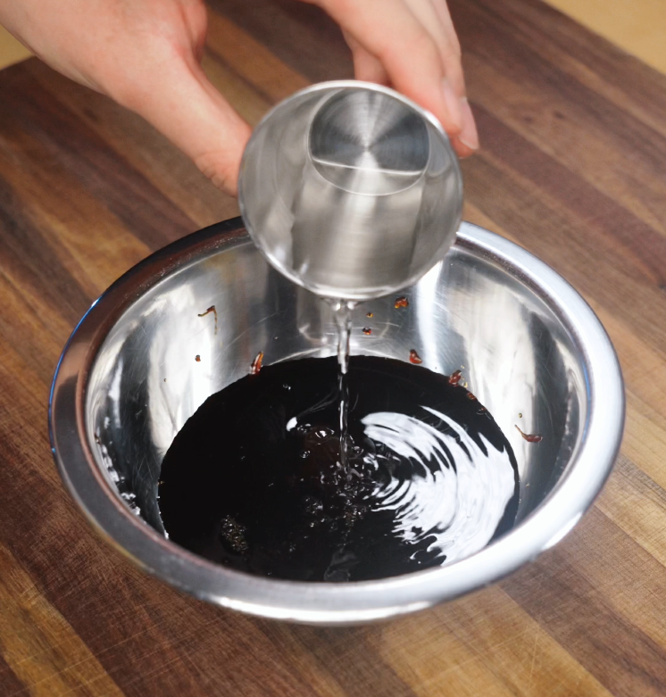 A stream of clear water is poured from a stainless steel cup into a metal bowl filled with dark soy-based sauce. This step is part of preparing the savory, tangy base for kung pao chicken, a classic Chinese stir-fry dish known for its bold flavors.
