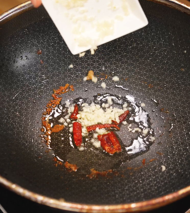 Minced garlic is added to a pan already sizzling with whole dried red chilies in hot oil. This step infuses the oil with heat and aroma, forming the spicy, fragrant foundation of kung pao chicken.