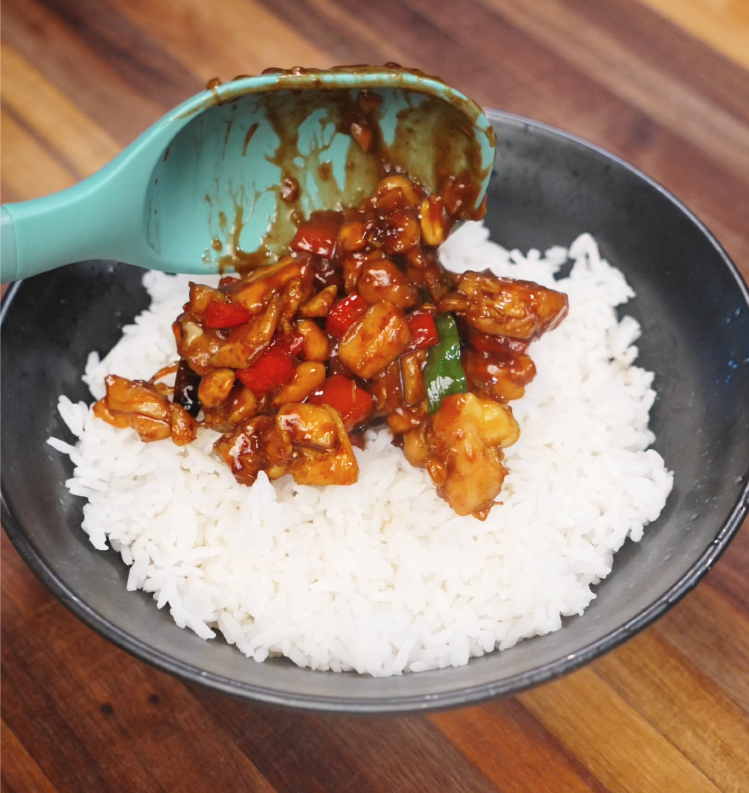 A ladle full of saucy kung pao chicken is served onto a bowl of freshly steamed white rice. The chicken, peanuts, and vegetables glisten with rich, savory sauce, making this a satisfying and colorful final dish.