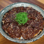 A bowl of Korean bulgogi beef served over white rice is topped with finely sliced scallions and toasted sesame seeds. The beef is glazed in a shiny sauce and arranged in a circular layer.