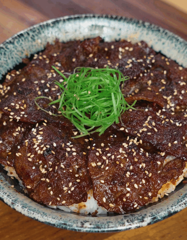 A bowl of Korean bulgogi beef served over white rice is topped with finely sliced scallions and toasted sesame seeds. The beef is glazed in a shiny sauce and arranged in a circular layer.
