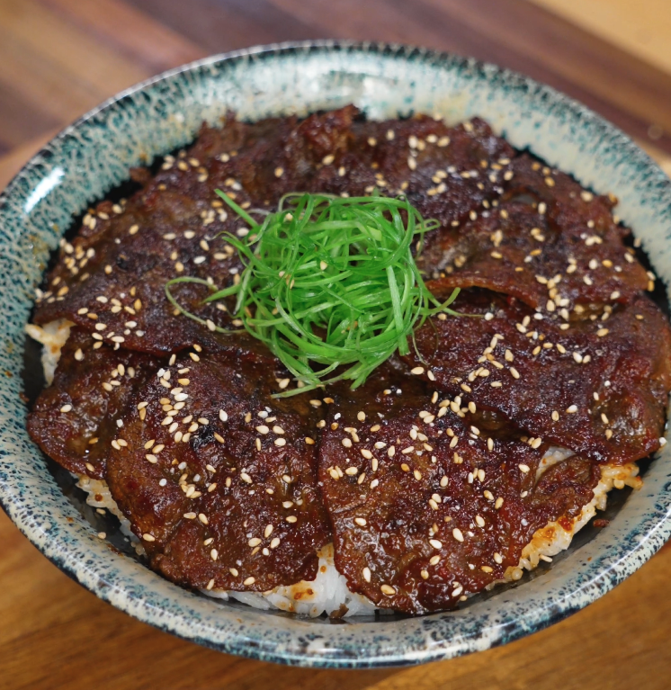 A bowl of Korean bulgogi beef served over white rice is topped with finely sliced scallions and toasted sesame seeds. The beef is glazed in a shiny sauce and arranged in a circular layer.