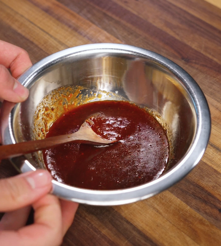 A wooden spoon is stirring a thick Korean bulgogi sauce inside a metal mixing bowl. The sauce has a rich reddish-brown color with a glossy texture, ready to coat the beef.