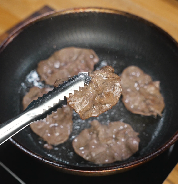 Slices of thinly cut beef are being seared in a hot pan, with tongs lifting one piece to show the browned surface. The meat is just beginning to caramelize for maximum flavor.