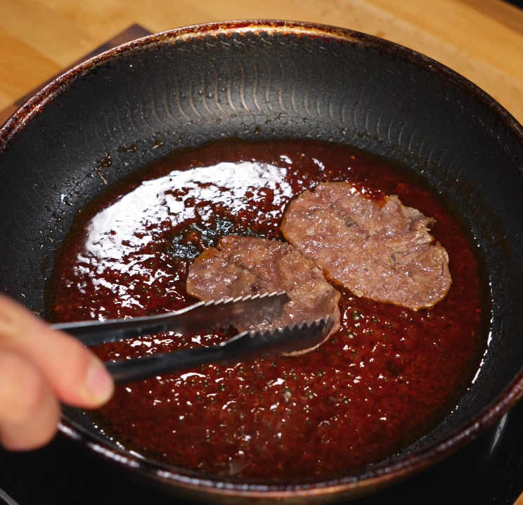 Thinly sliced beef is simmering in a bubbling sweet and savory bulgogi-style sauce in a skillet. The glossy sauce thickens around the meat, infusing it with deep umami flavor.