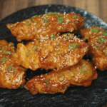 Plated Korean fried chicken tenders topped with sesame seeds and sliced scallions, ready to serve on a black ceramic plate.