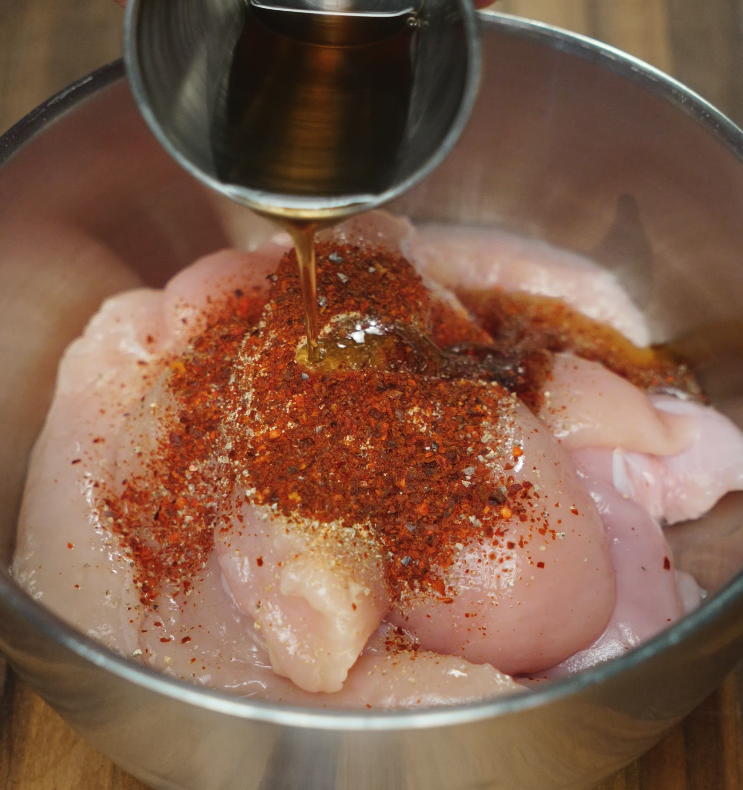 Pouring marinade over raw chicken tenderloins in a metal bowl, showing the start of the Korean fried chicken preparation with spices and oil.