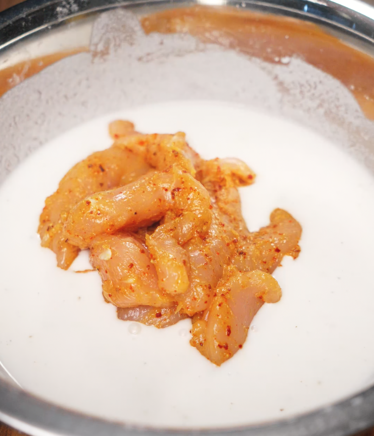 Marinated chicken tenders coated in a creamy buttermilk-based batter in a silver mixing bowl, ready for frying.
