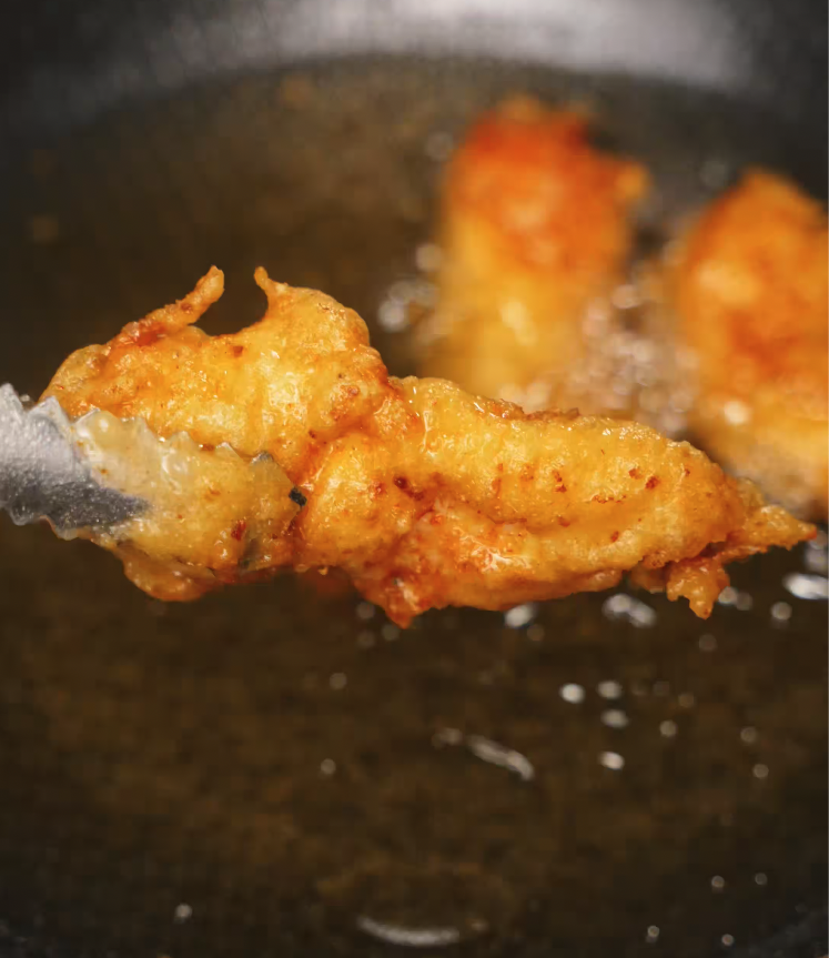 Freshly battered chicken tender being lifted from hot oil with tongs, showing golden brown, crispy Korean-style breading.
