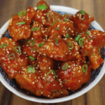 Korean fried chicken coated in a glossy red sauce, garnished with sesame seeds and chopped scallions in a bowl.