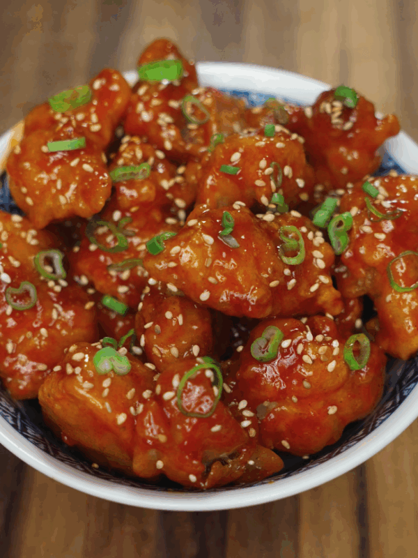 Korean fried chicken coated in a glossy red sauce, garnished with sesame seeds and chopped scallions in a bowl.