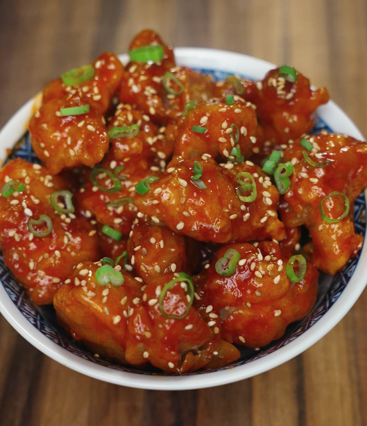 Korean fried chicken coated in a glossy red sauce, garnished with sesame seeds and chopped scallions in a bowl.
