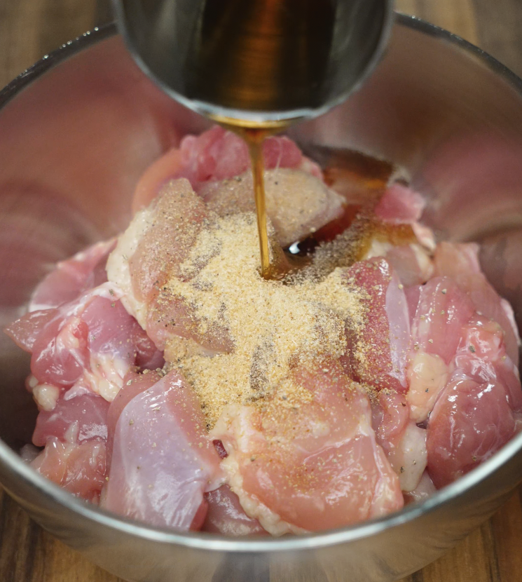 Soy sauce being poured over raw chicken with garlic powder in a metal mixing bowl for marinating.