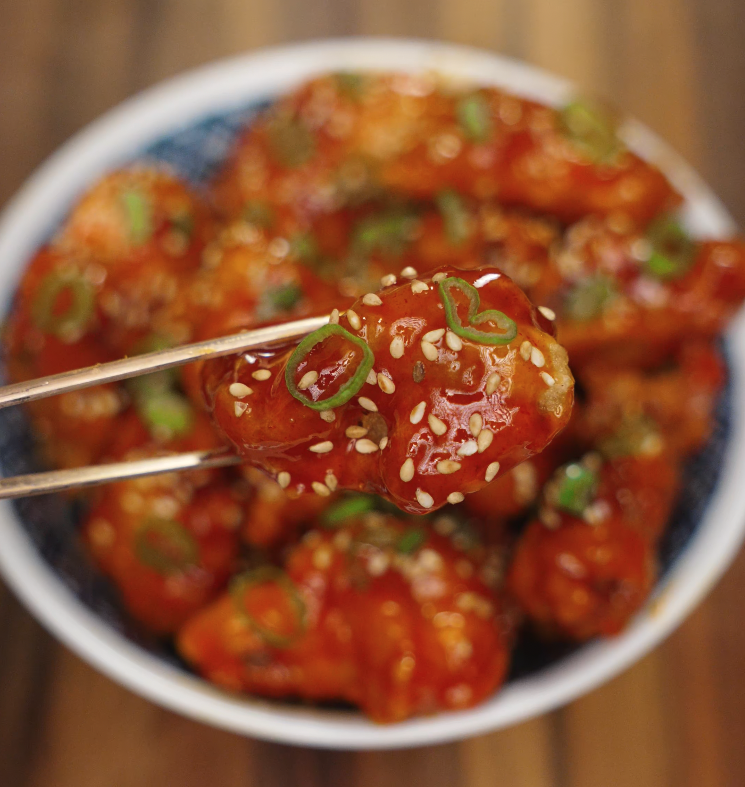 A single piece of spicy Korean fried chicken garnished with sesame seeds and scallions being held with chopsticks.