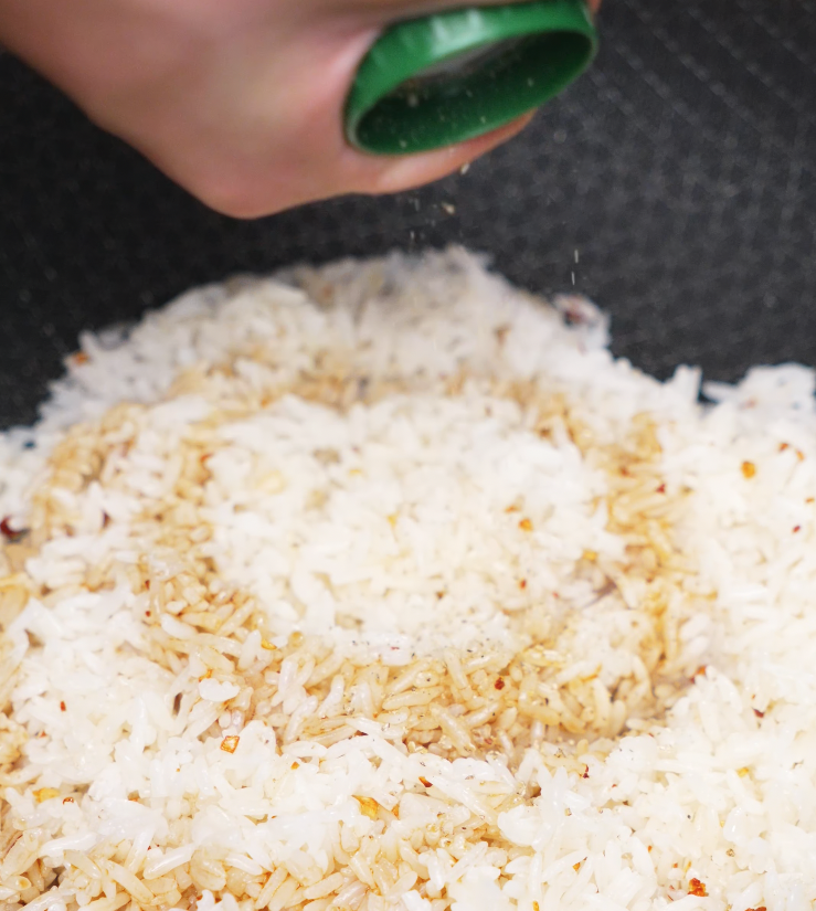 A close-up shot of white jasmine rice being seasoned with salt and spices in a non-stick frying pan as part of the garlic butter fried rice cooking process. The rice is slightly golden from the garlic-infused butter and shows hints of red pepper flakes for flavor. This step is essential for creating a fragrant and flavorful base for garlic butter fried rice, a popular and easy Asian-inspired weeknight dinner recipe.