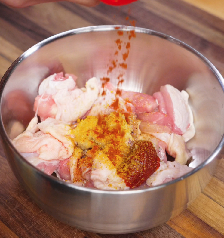A hand sprinkles red chili powder over raw boneless chicken thighs in a bowl with ginger paste, garlic, and a dollop of gochujang. The bold ingredients hint at a spicy, savory marinade that will give the chicken deep flavor and color.