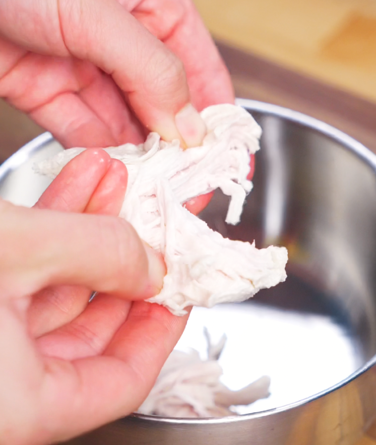 Hands gently shredding freshly poached chicken breast into fine, tender strands over a metal mixing bowl. This shredded chicken will be tossed with peanut sauce and noodles to create a protein-packed, creamy Asian-style noodle dish.