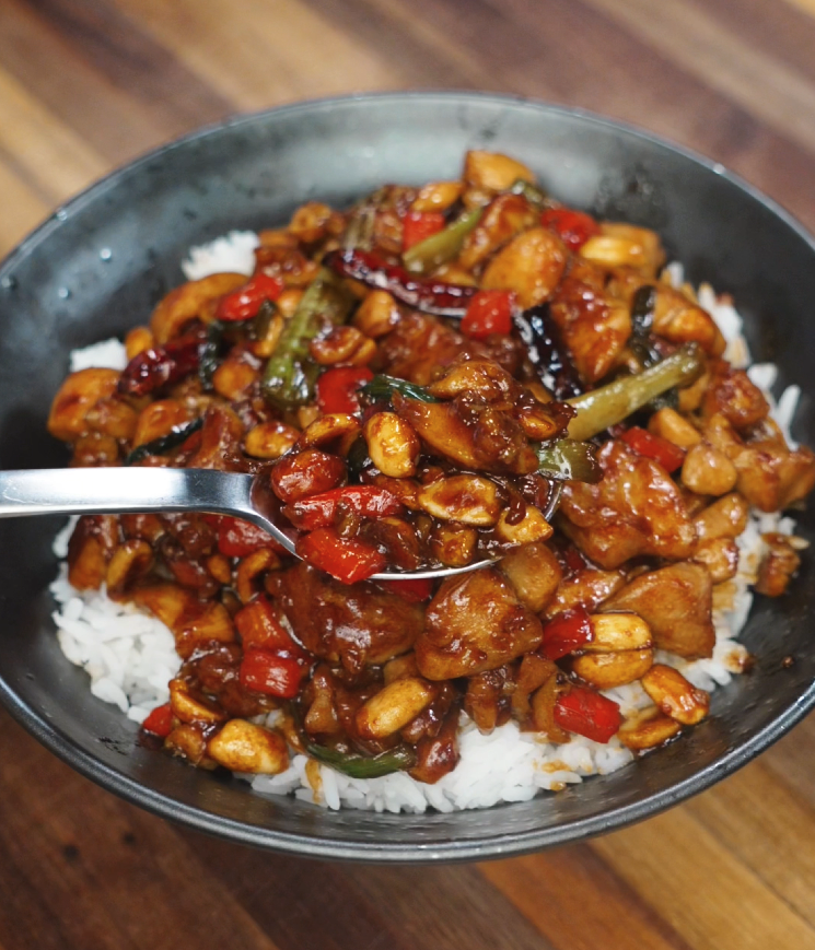 A large spoon lifts a portion of kung pao chicken from the bowl, showing saucy chunks of stir-fried chicken, peanuts, red peppers, and scallions. The glossy sauce clings to every ingredient, ready to be enjoyed over rice.