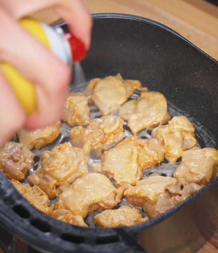 A hand sprays cooking oil over evenly spaced marinated chicken thigh pieces inside an air fryer basket. The coating ensures the chicken crisps up during cooking while retaining its juicy interior.