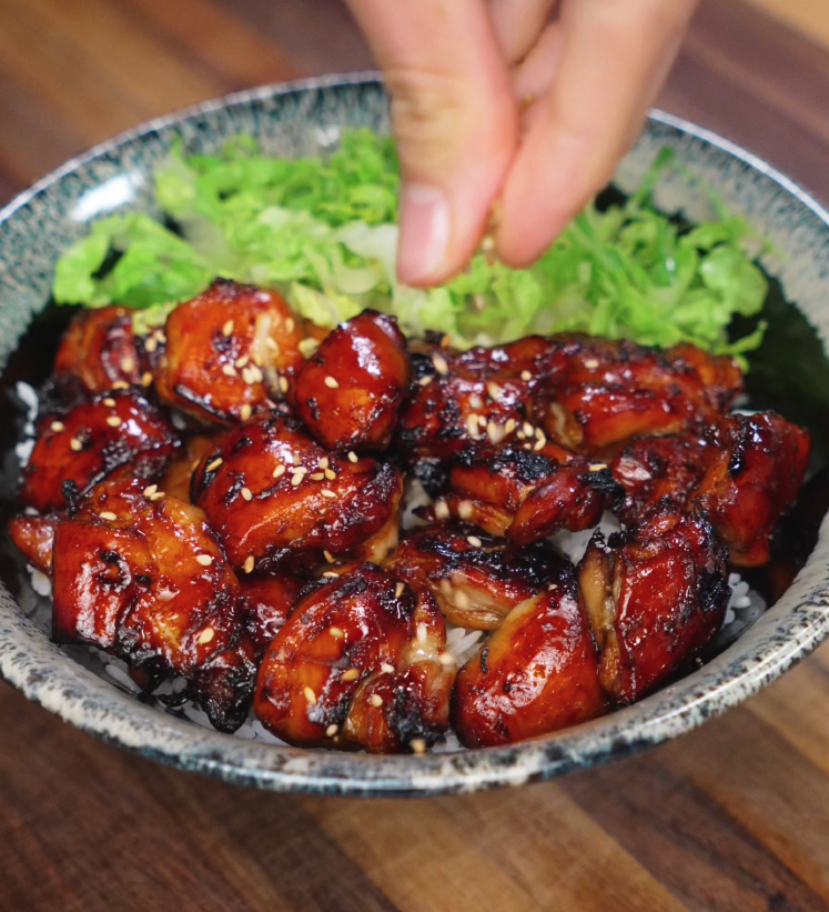 A hand sprinkles sesame seeds over a freshly cooked bowl of garlic soy glazed chicken. The chicken pieces are deeply caramelized with crisp edges and glistening glaze, sitting atop white rice with a side of crisp lettuce, creating the perfect final touch for texture and visual appeal.