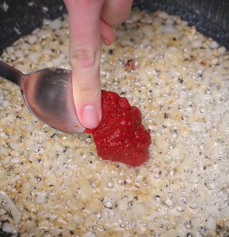 A spoon with tomato paste is being pressed into a pan of sizzling sautéed onions, starting the rich base for the Marry Me Chicken pasta sauce.