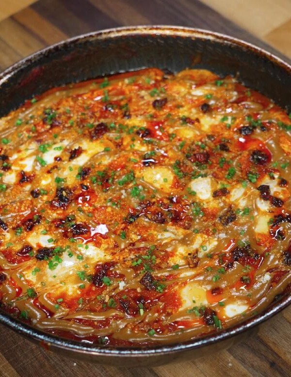 The completed peanut chili oil shakshuka served in the skillet, topped with drizzles of sauce, crispy chili oil, and finely chopped herbs.