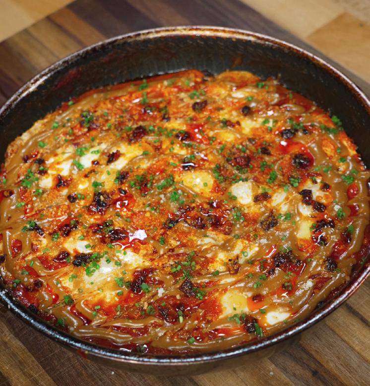 The completed peanut chili oil shakshuka served in the skillet, topped with drizzles of sauce, crispy chili oil, and finely chopped herbs.