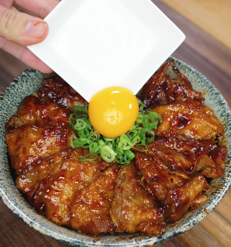 Carefully placing a raw egg yolk on top of chopped green onions and sticky spicy pork belly slices arranged in a rice bowl, highlighting the contrast in textures and vibrant colors.