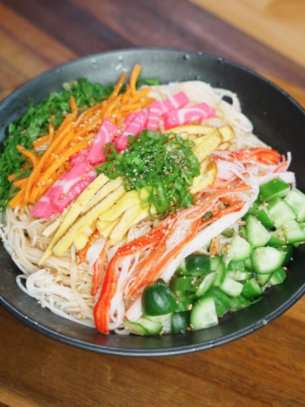 A fully assembled somen salad bowl featuring chilled noodles topped with julienned egg, imitation crab, cucumber, carrots, pink fish cake, and scallions with sesame seeds sprinkled on top.