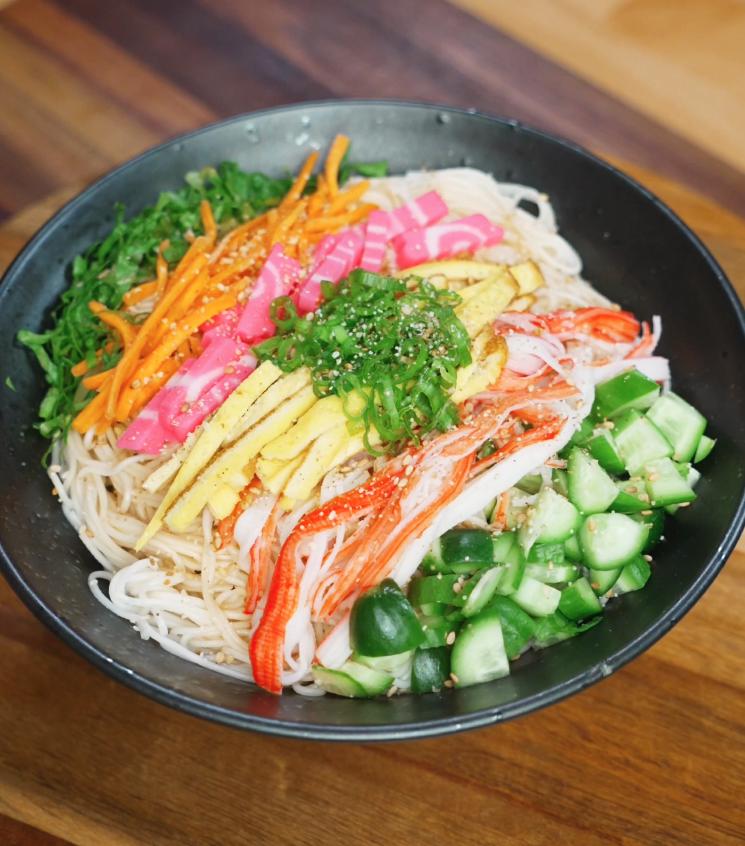 A fully assembled somen salad bowl featuring chilled noodles topped with julienned egg, imitation crab, cucumber, carrots, pink fish cake, and scallions with sesame seeds sprinkled on top.