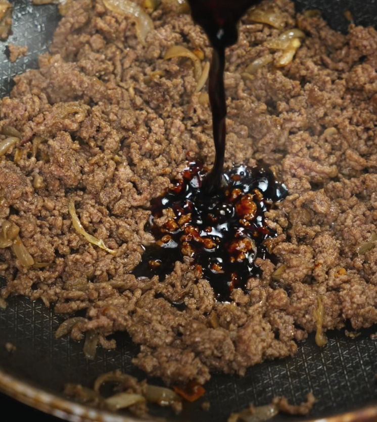 A dark, glossy sweet-savory Thai sauce is being poured over cooked ground beef and onions, infusing it with rich umami flavor.