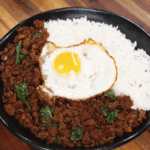 A black bowl filled with fragrant pad kra pao topped with crispy Thai basil, steamed white rice, and a perfectly fried sunny-side-up egg.