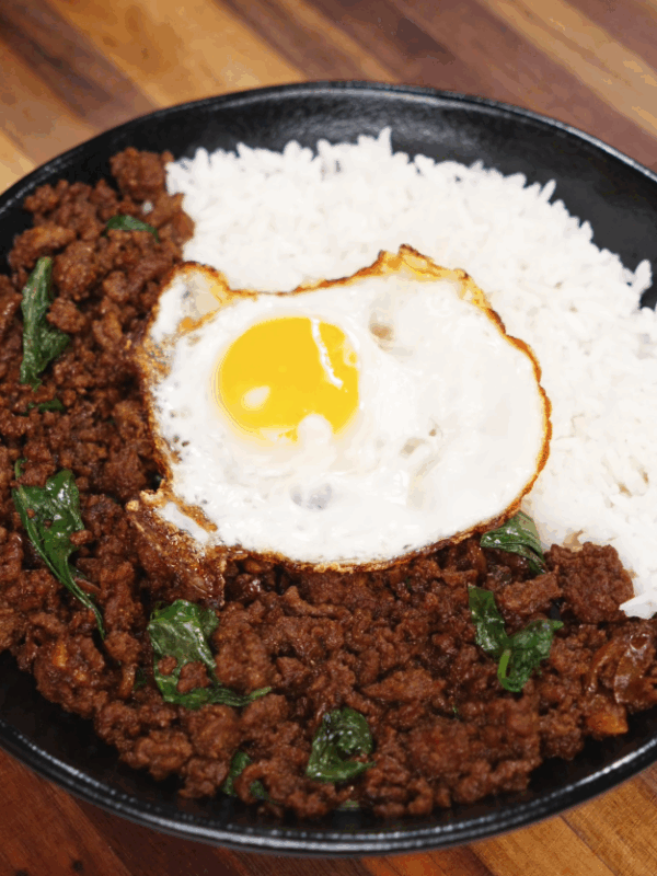 A black bowl filled with fragrant pad kra pao topped with crispy Thai basil, steamed white rice, and a perfectly fried sunny-side-up egg.