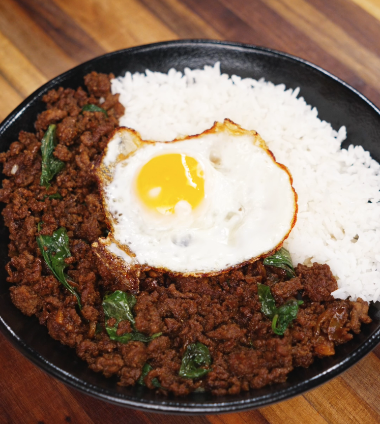 A black bowl filled with fragrant pad kra pao topped with crispy Thai basil, steamed white rice, and a perfectly fried sunny-side-up egg.