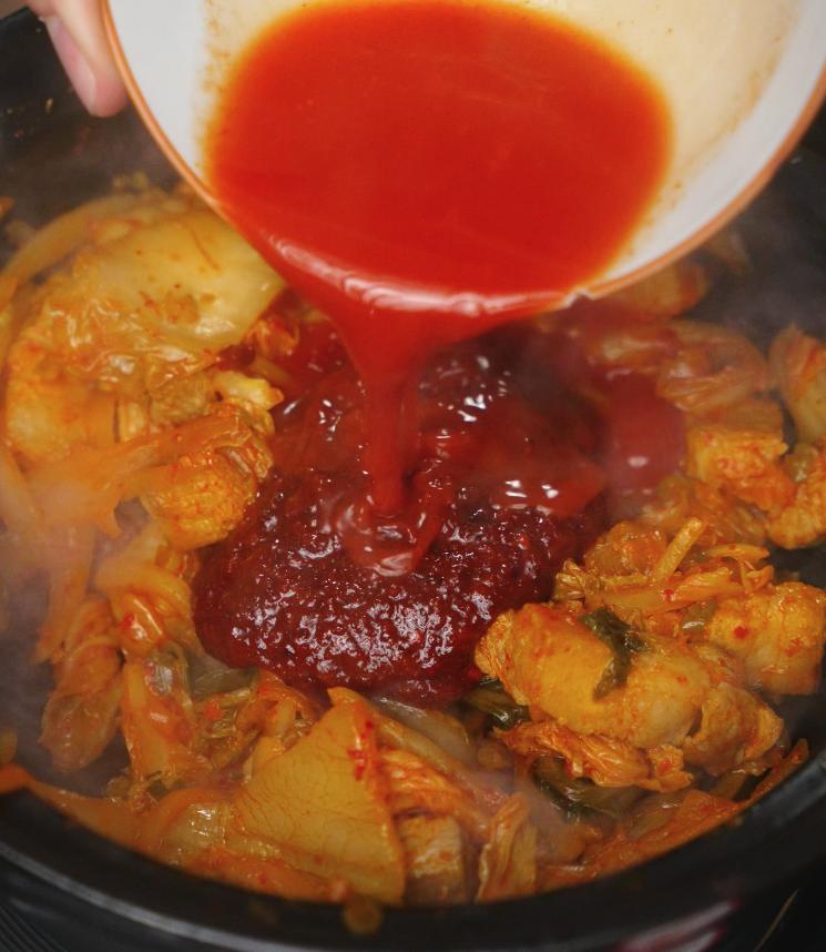 Pouring a vibrant red kimchi soup base into a simmering pot filled with pork belly and napa cabbage. The gochugaru-infused sauce immediately blends with the bubbling stew, enhancing the spicy, savory aroma of the dish.