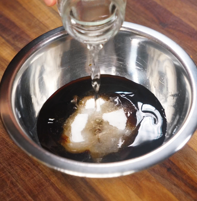 Adding rice vinegar to a homemade stir fry sauce mixture of soy sauce, sugar, and aromatics in a metal bowl on a wooden surface.