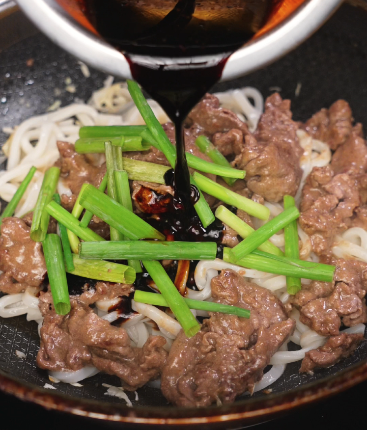 Pouring black pepper sauce over stir-fried beef, scallions, and udon noodles in a skillet during the final cooking step.