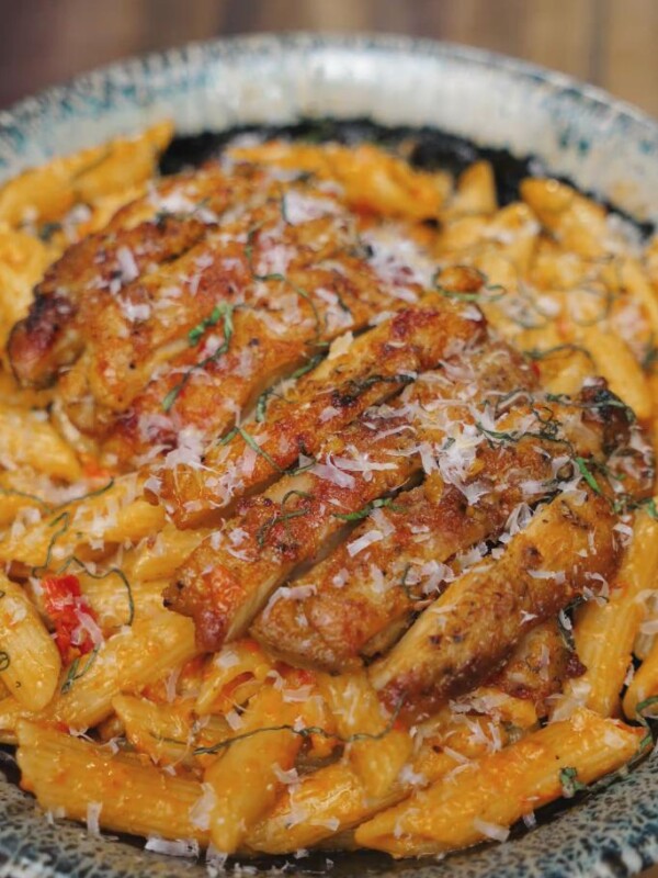 A bowl of penne pasta smothered in creamy tomato sauce is topped with sliced seared chicken and freshly grated parmesan in the Marry Me Chicken pasta dish.