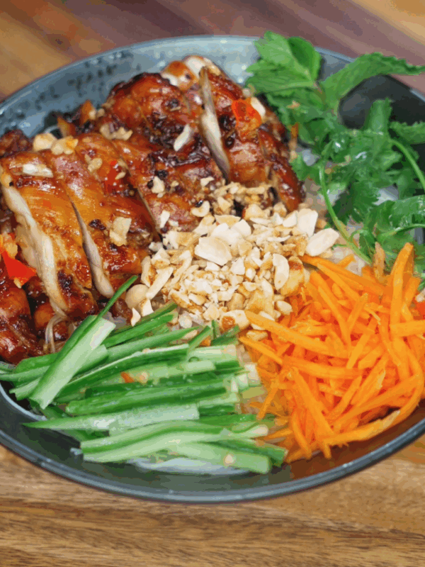 Fully assembled Vietnamese lemongrass chicken vermicelli bowl with grilled chicken, crushed peanuts, julienned carrots, cucumbers, mint, and cilantro on top of rice noodles.