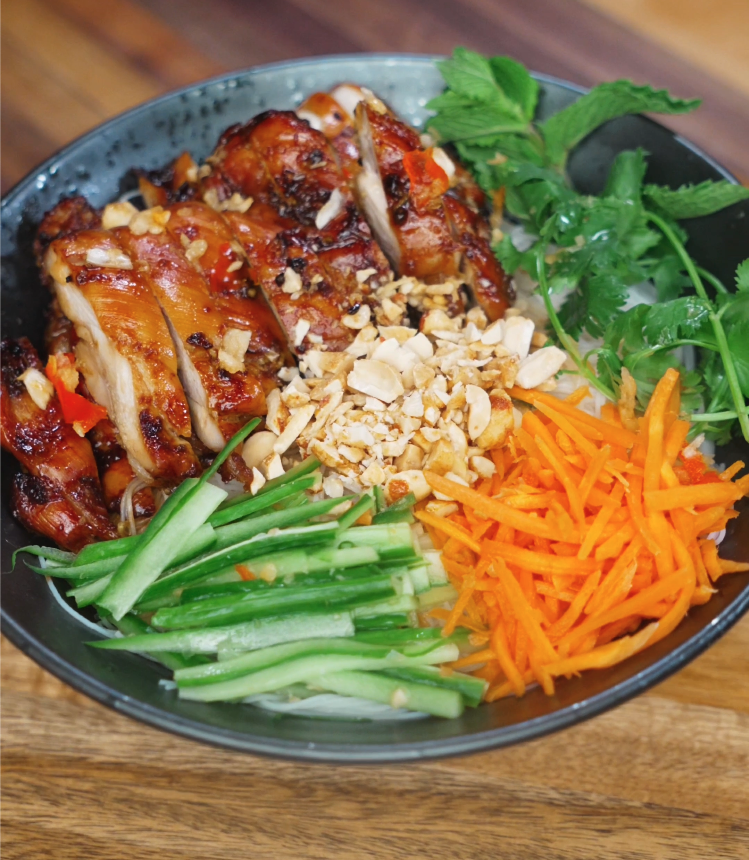 Fully assembled Vietnamese lemongrass chicken vermicelli bowl with grilled chicken, crushed peanuts, julienned carrots, cucumbers, mint, and cilantro on top of rice noodles.