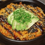 Finished sushi bake topped with creamy avocado slices, scallions, sesame seeds, and a drizzle of unagi sauce in a black ceramic baking dish.