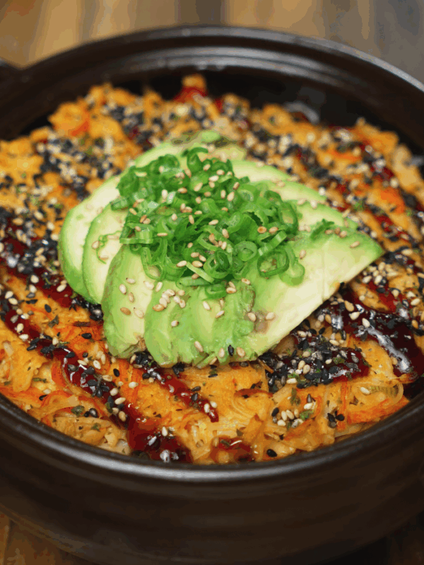 Finished sushi bake topped with creamy avocado slices, scallions, sesame seeds, and a drizzle of unagi sauce in a black ceramic baking dish.