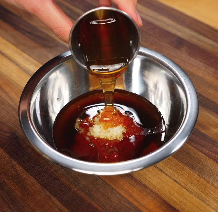 A hand pours golden honey into a stainless steel bowl filled with grated garlic, soy sauce, and other marinade ingredients, creating a glossy and flavorful sweet-savory base for the jjolmyeon noodles.