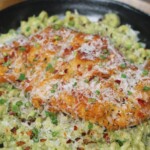 Close-up of golden, crunchy chicken breast resting on a bed of creamy pesto fusilli, garnished with parmesan and fresh green herbs.